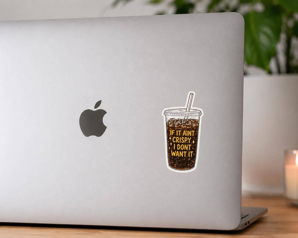 A silver laptop with an Apple logo features the Funny Crispy Diet Coke sticker - Matte Vinyl by Hello Brand | Sticker My Art. A plant and candle are blurred in the background.