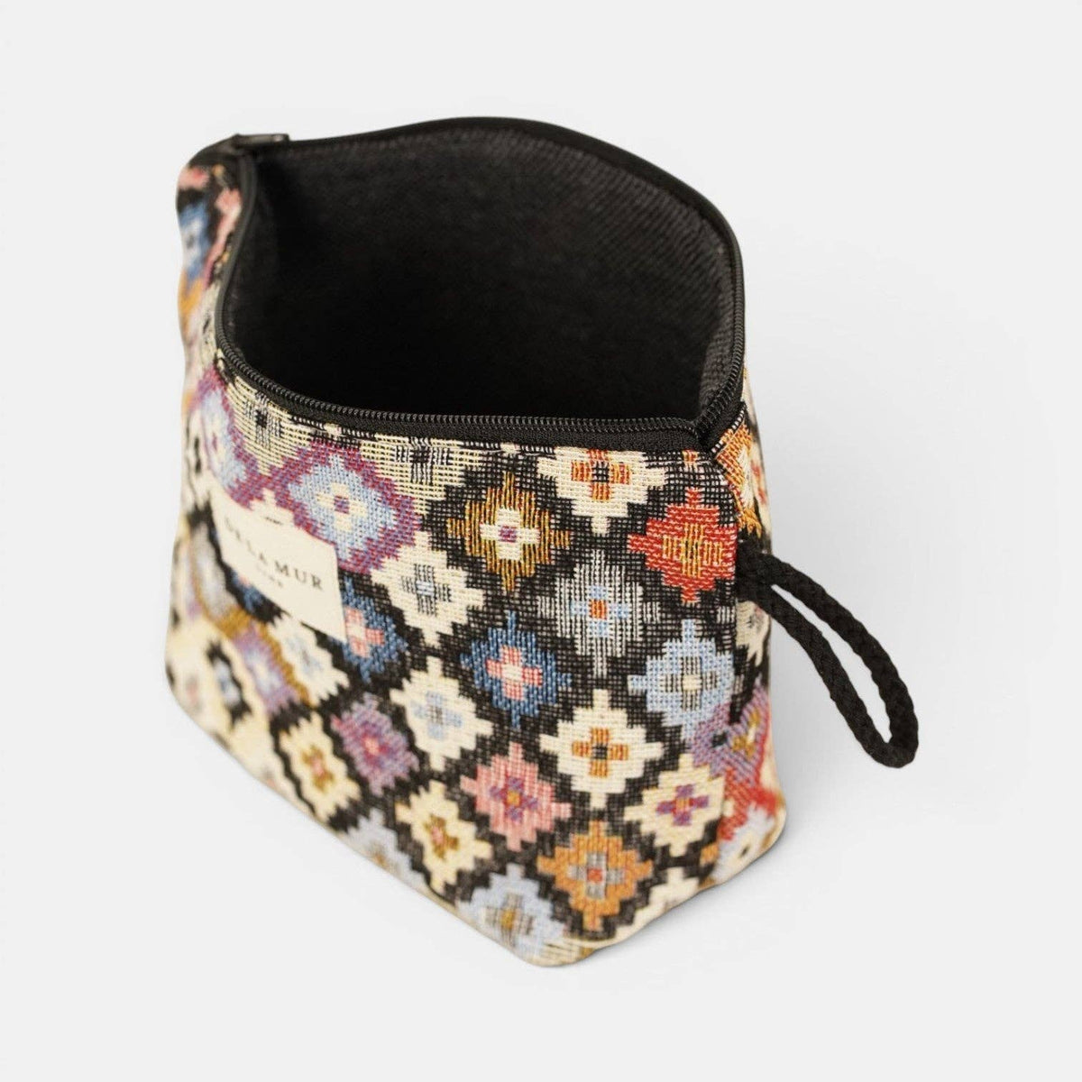 The Geometric Jacquard Small Makeup Bag – Multicolor (Ore) by DE LA MUR LINE US Inc is a compact makeup bag made from jacquard fabric, showcasing a vibrant geometric diamond pattern. It features a black zipper and matching black side loop, and is displayed standing open on a plain white background.