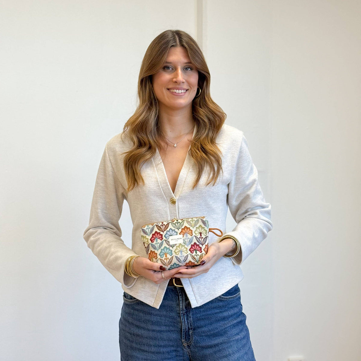 A woman with long brown hair, dressed in a light sweater and blue jeans, smiles as she holds a decorative mug and the DE LA MUR LINE US Inc Floral Jacquard Small Makeup Bag – Multicolor (Flora), standing against a plain white background.
