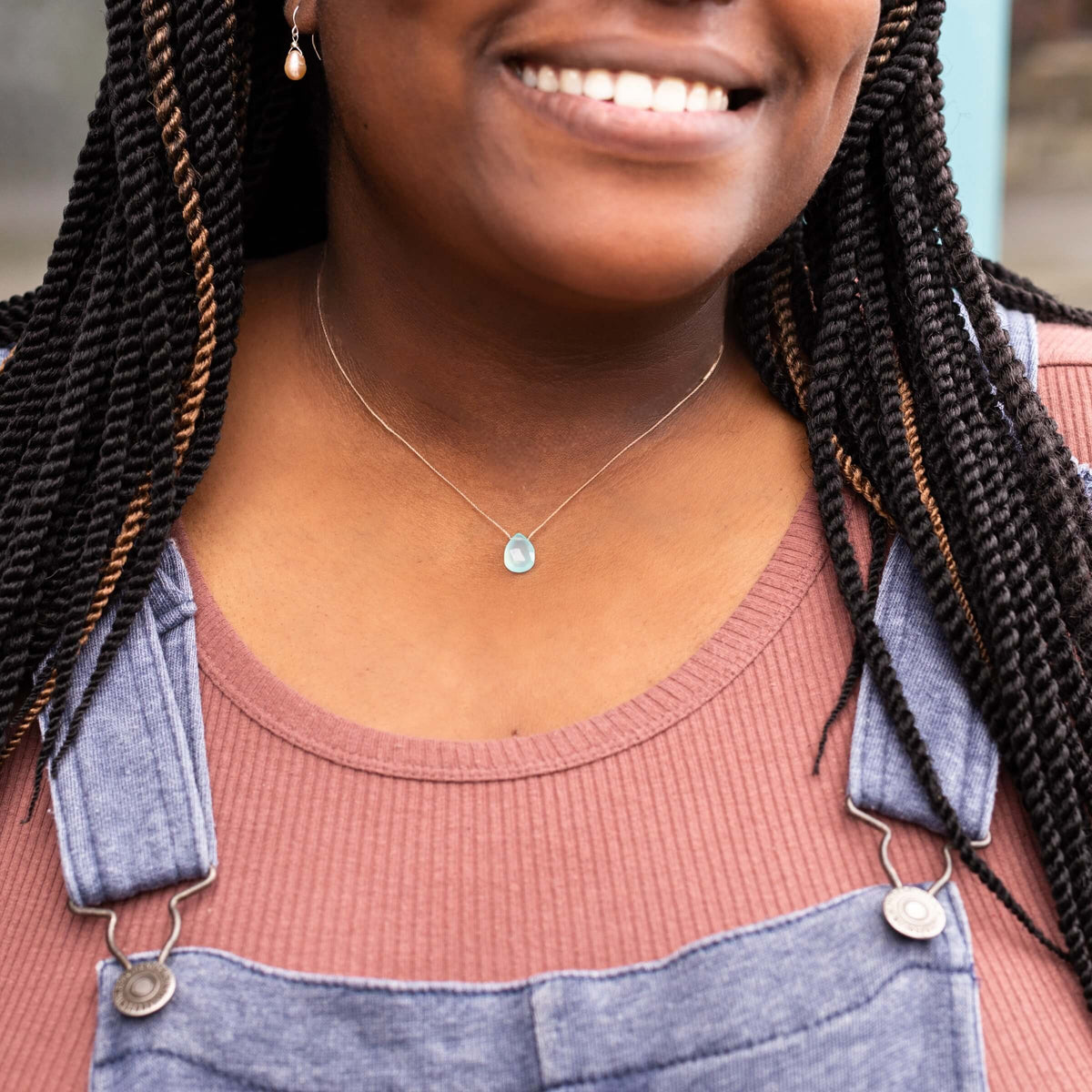 A smiling person in a pink top and denim overalls wears handmade gemstone jewelry—SoulKu’s Chalcedony Luxe Necklace for Happy Birthday—and a single pearl earring. The image is cropped to show only the lower face and torso.