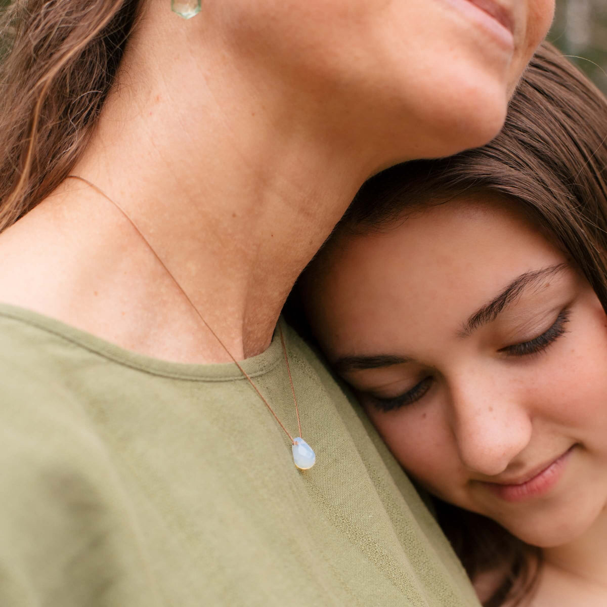 A young girl rests her head on a woman’s shoulder. The woman, looking content in a green top, wears the Opaline Crystal Necklace Gift for Motherhood - Mom, Mother’s by SoulKu.