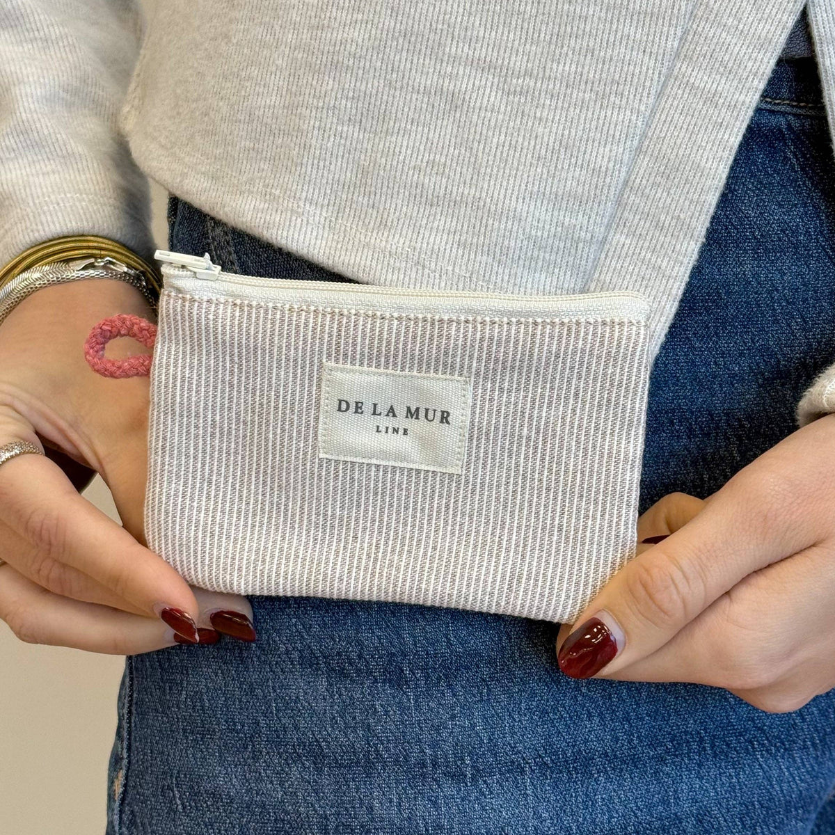 A person dressed in blue jeans and a light sweater holds a small, striped coin purse in gray and cream from DE LA MUR LINE US Inc. The person has rings on their fingers and a pink flower tattoo on their hand.