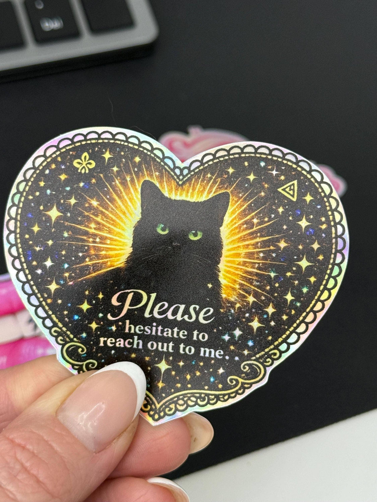A hand holds the Black Cat Holographic “Please Hesitate to Reach Out” Sticker by Hello Brand | Sticker My Art, featuring a heart-shaped black cat design with glowing eyes and text: Please hesitate to reach out to me.