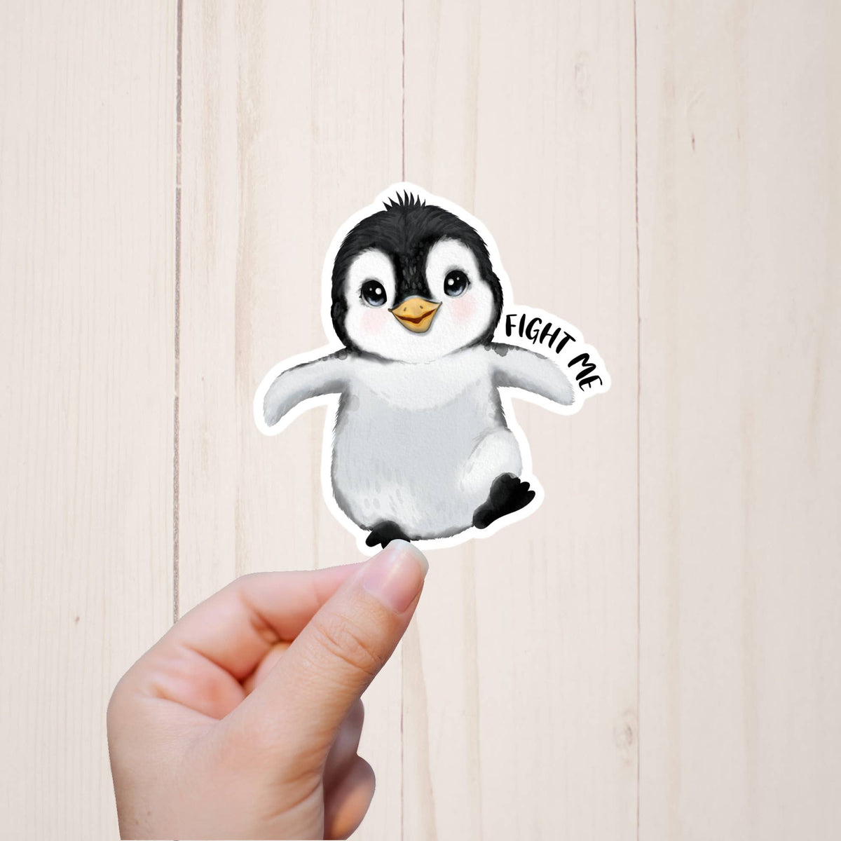 A hand holds a 3″ x 1.5″ Penguin Fight Me Sticker by Sorry Not Sorry Co, showing a cute baby penguin and bold text against a light wooden background.