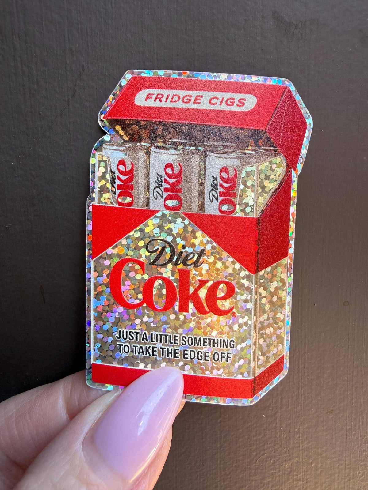 A hand holds the Diet Coke Fridge Cigs Sticker by Hello Brand | Sticker My Art—a glittery vinyl sticker shaped like a cigarette pack filled with mini Diet Coke cans. Weather-resistant.