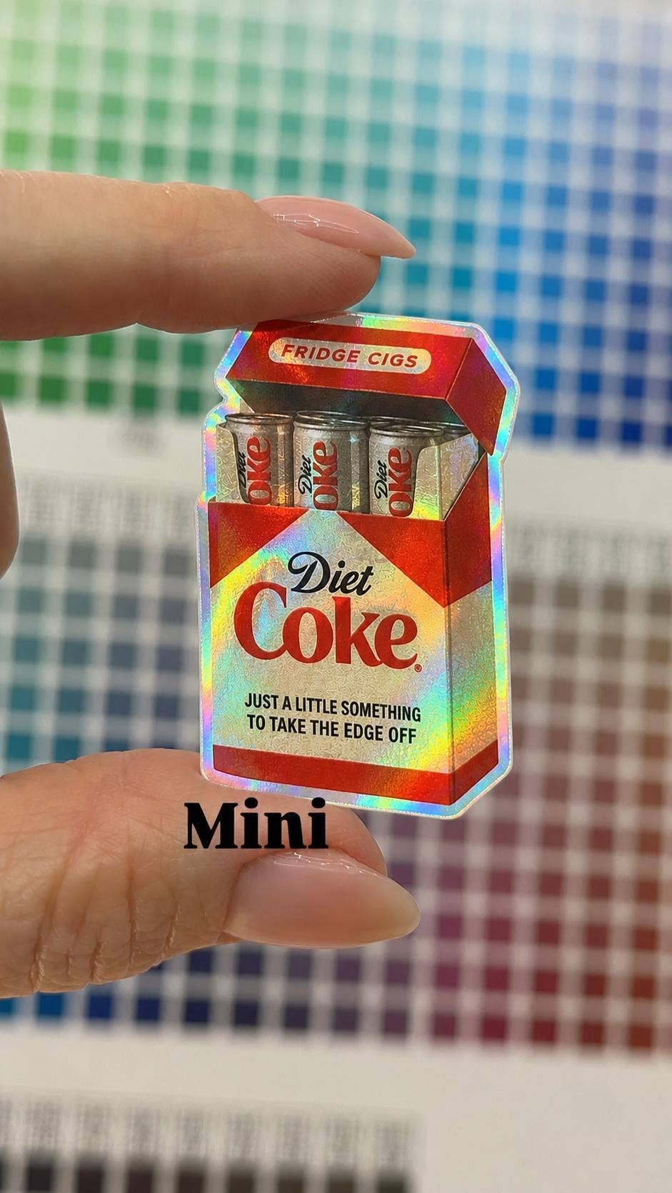 A hand holds a weather-resistant Diet Coke Fridge Cigs Sticker by Hello Brand | Sticker My Art, featuring a pack of cans arranged like cigarettes and captioned, Just a little something to take the edge off.