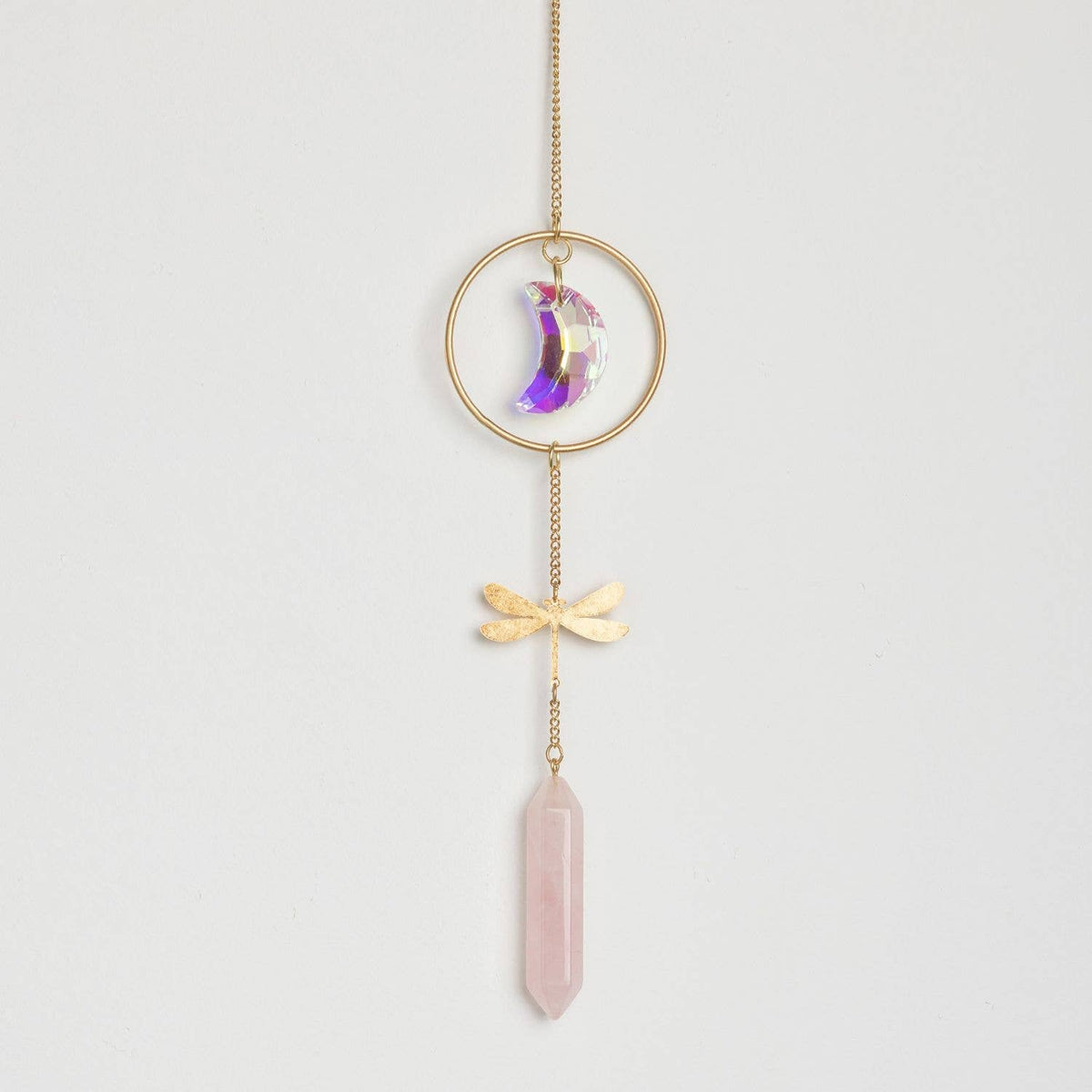 Introducing the Stone Suncatcher - Dragonfly/Rose Quartz from Scout Curated Wears: a stunning window ornament featuring a gold ring with an iridescent crescent moon, accented by a delicate gold dragonfly charm and finished with a rose quartz crystal pendant. All elements are artfully suspended from a gold chain, creating an elegant display against any backdrop. This versatile piece not only elevates your décor but also catches sunlight to cast gentle reflections throughout the room.