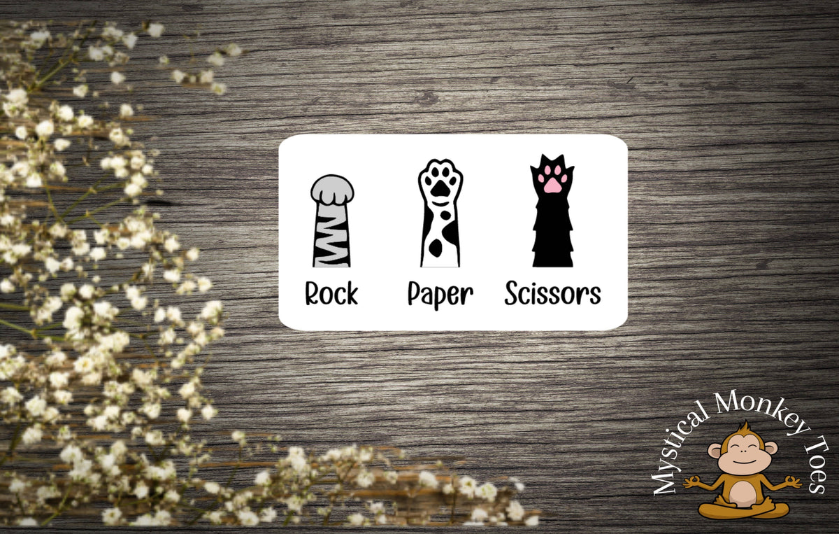 A card with cartoon animal limbs for Rock Paper Scissors sits on wood. The Mystical Monkey Toes logo is at the bottom right, with white flowers and a Cat Paws Sticker in waterproof vinyl on the left.