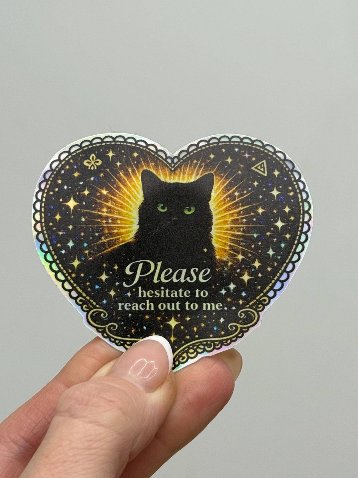 A hand holds the Black Cat Holographic “Please Hesitate to Reach Out” Sticker from Hello Brand | Sticker My Art, featuring a black cat with green eyes, glowing rays, stars, and humorous text.
