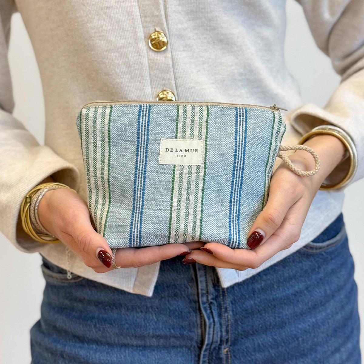A person in a cream cardigan and blue jeans holds the Striped Jacquard Small Makeup Bag – Blue & Green (Cata) by DE LA MUR LINE US Inc. Their nails are painted red, and they wear gold bracelets.