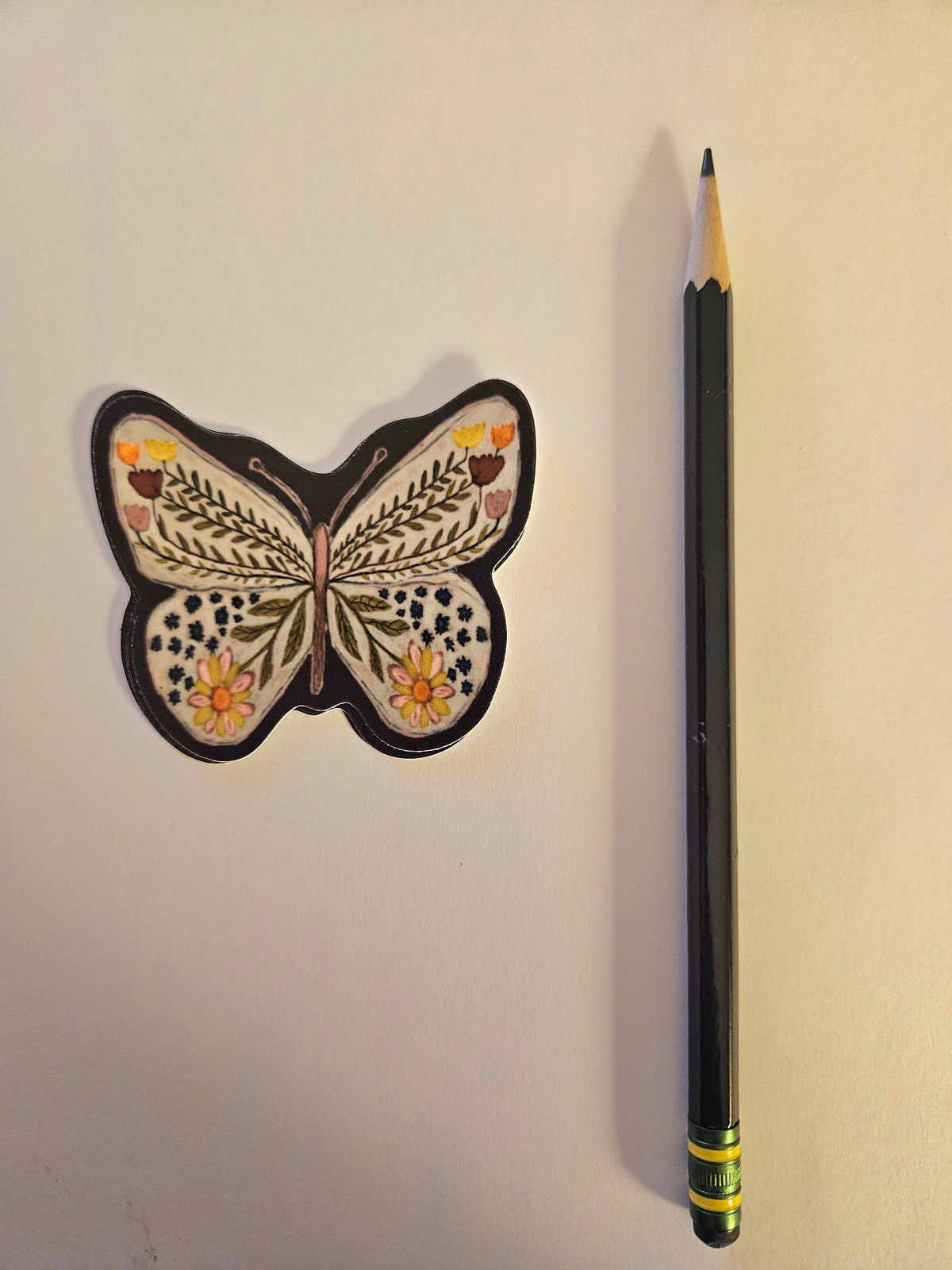 A pencil with an eraser sits beside the Butterfly & Flowers Vinyl Sticker by Blueberry Road Cards, featuring yellow, brown, green, and white floral and leaf designs on a plain light surface.