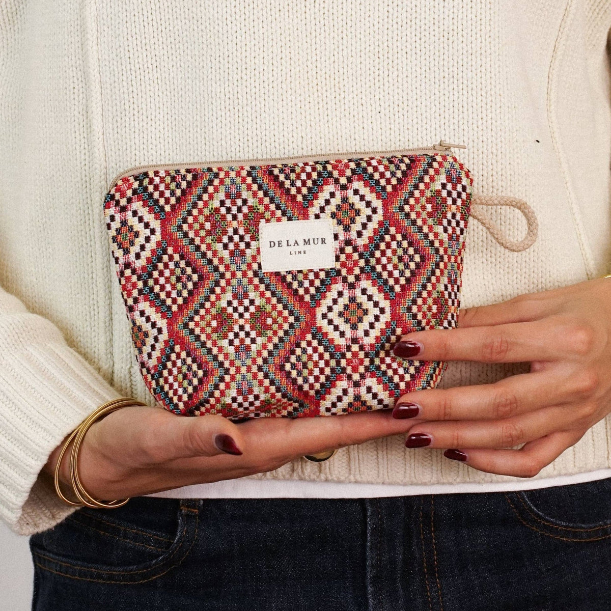 A person in a cream sweater and dark jeans holds the DE LA MUR LINE US Inc “Geometric Jacquard Small Makeup Bag – Red Multicolor (Otto)” with both hands. The bag features a bold red, pink, and white geometric jacquard pattern and is adorned with a small white label.