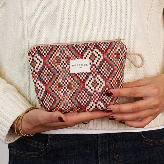 A person in a cream sweater and dark jeans holds the DE LA MUR LINE US Inc “Geometric Jacquard Small Makeup Bag – Red Multicolor (Otto)” with both hands. The bag features a bold red, pink, and white geometric jacquard pattern and is adorned with a small white label.