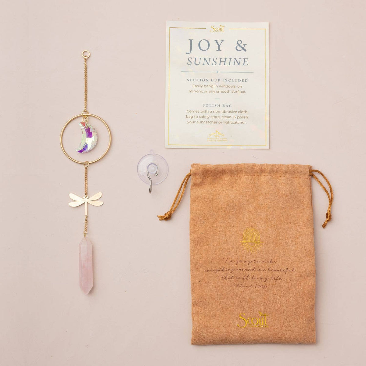 The Scout Curated Wears Stone Suncatcher - Dragonfly/Rose Quartz, featuring a pink crystal window hanging, comes with a suction cup, a tan storage pouch, and a Joy & Sunshine card—all displayed on a beige background.