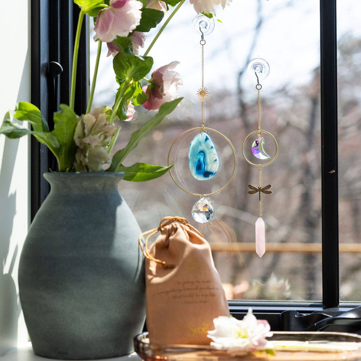 A vase with pink and white flowers sits by a window. Hanging from the glass is the Scout Curated Wears Stone Suncatcher in the Sun Rays/Black/Blue Agate style, featuring unique crystals and gold details that catch the sunlight. Nearby on the windowsill rests a tan drawstring bag.