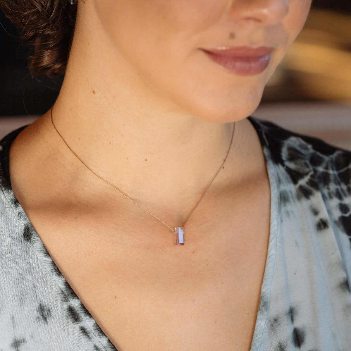 A woman wears the SoulKu Amethyst Refined Necklace for Fearless—a delicate handmade necklace featuring a small rectangular amethyst pendant—with a light blue patterned top, shown from the shoulders up.