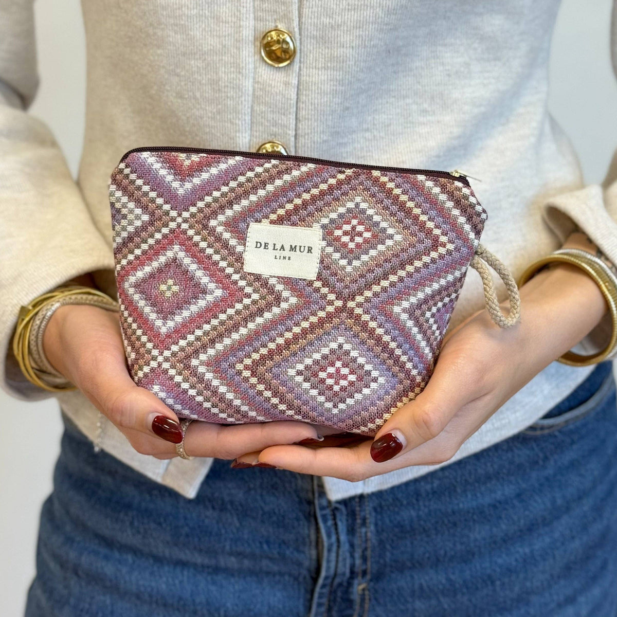 A person in a beige sweater and blue jeans holds the Geometric Jacquard Small Makeup Bag – Pink & Lilac (Nana) by DE LA MUR LINE US Inc. The makeup bag features a striking geometric kilim pattern in shades of pink and lilac, with the DE LA MUR label visible.