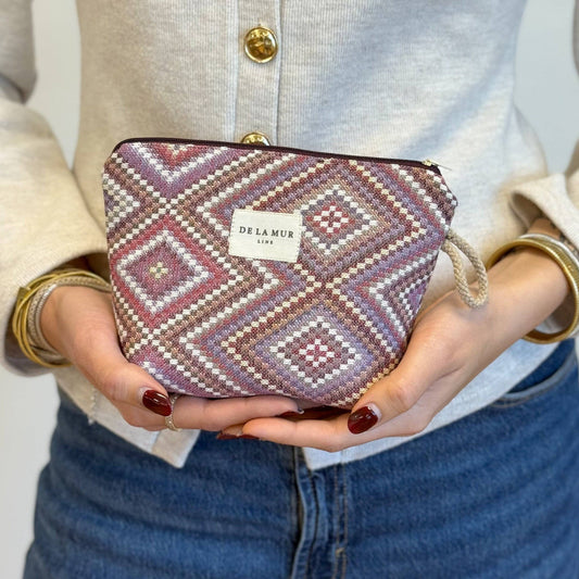 A person in a beige sweater and blue jeans holds the Geometric Jacquard Small Makeup Bag – Pink & Lilac (Nana) by DE LA MUR LINE US Inc. The makeup bag features a striking geometric kilim pattern in shades of pink and lilac, with the DE LA MUR label visible.