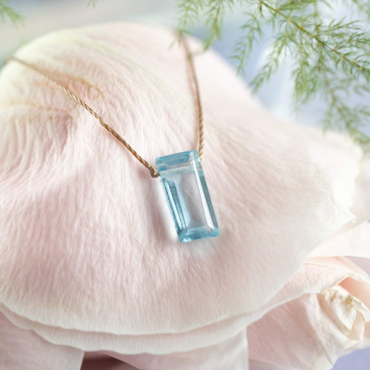 The Sky Blue Topaz Refined Necklace for New Beginnings by SoulKu features a rectangular pendant on a slender chain, elegantly displayed on pink seashells with soft green foliage in the background.