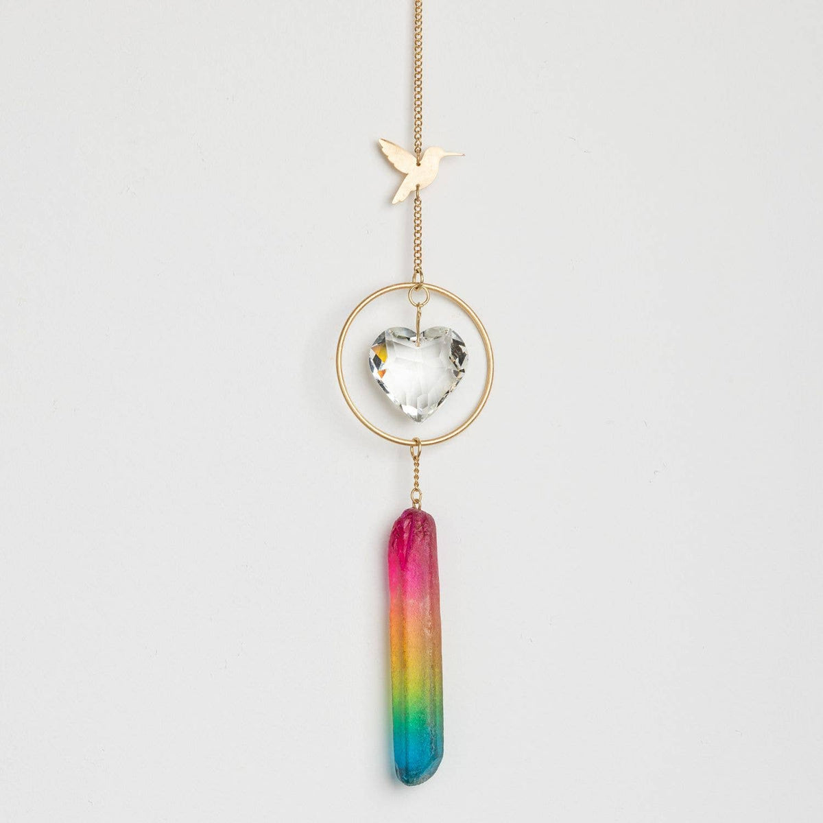 The Stone Suncatcher - Hummingbird/Rainbow Stone by Scout Curated Wears features a gold chain adorned with a small hummingbird charm, a clear crystal heart encircled in gold, and a dangling rainbow-colored crystal wand reminiscent of a suncatcher, all showcased against a plain white background.