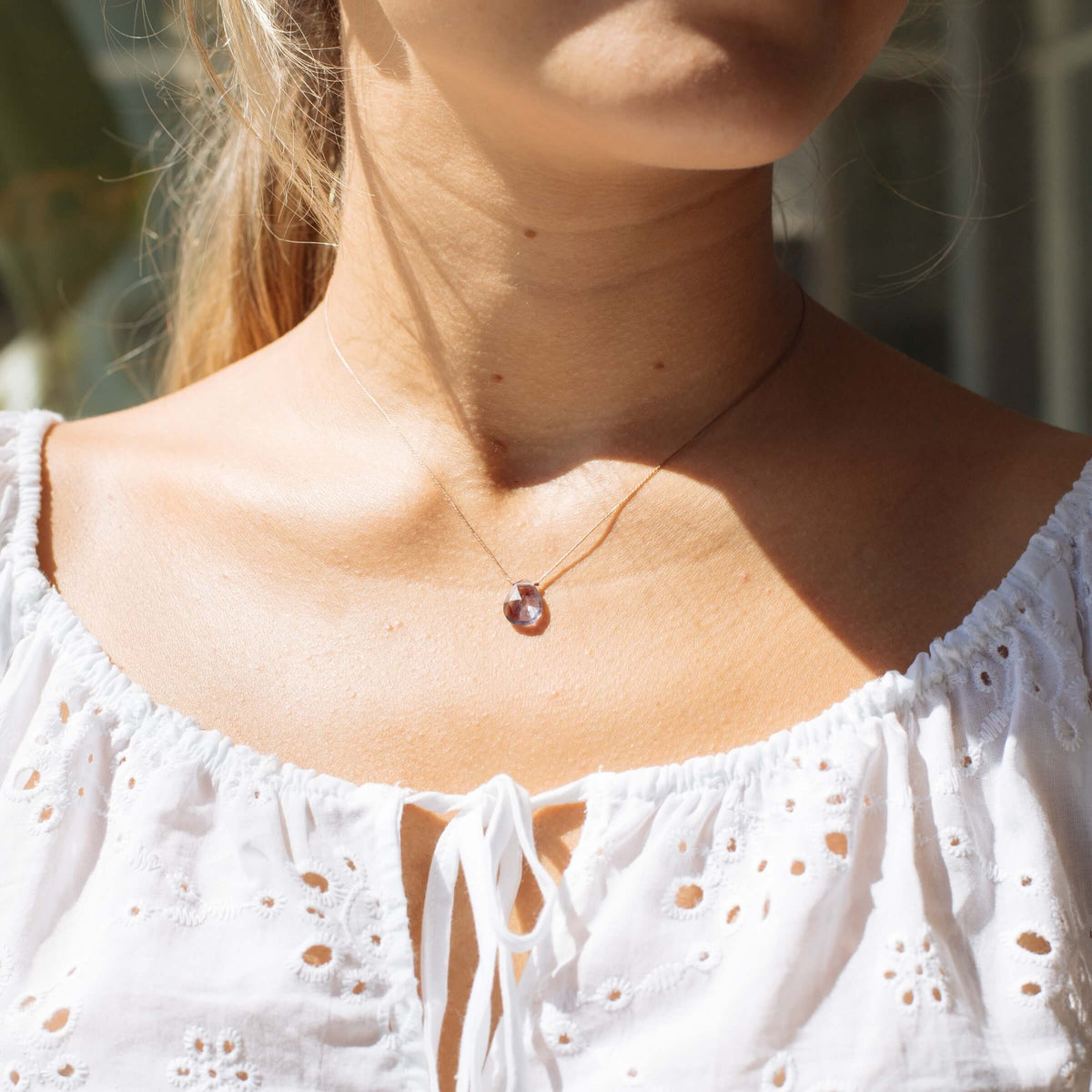 A woman in a white eyelet blouse wears the Fluorite Luxe Necklace for Magic by SoulKu, sunlight highlighting her neck and collarbone while her face is partially out of frame.
