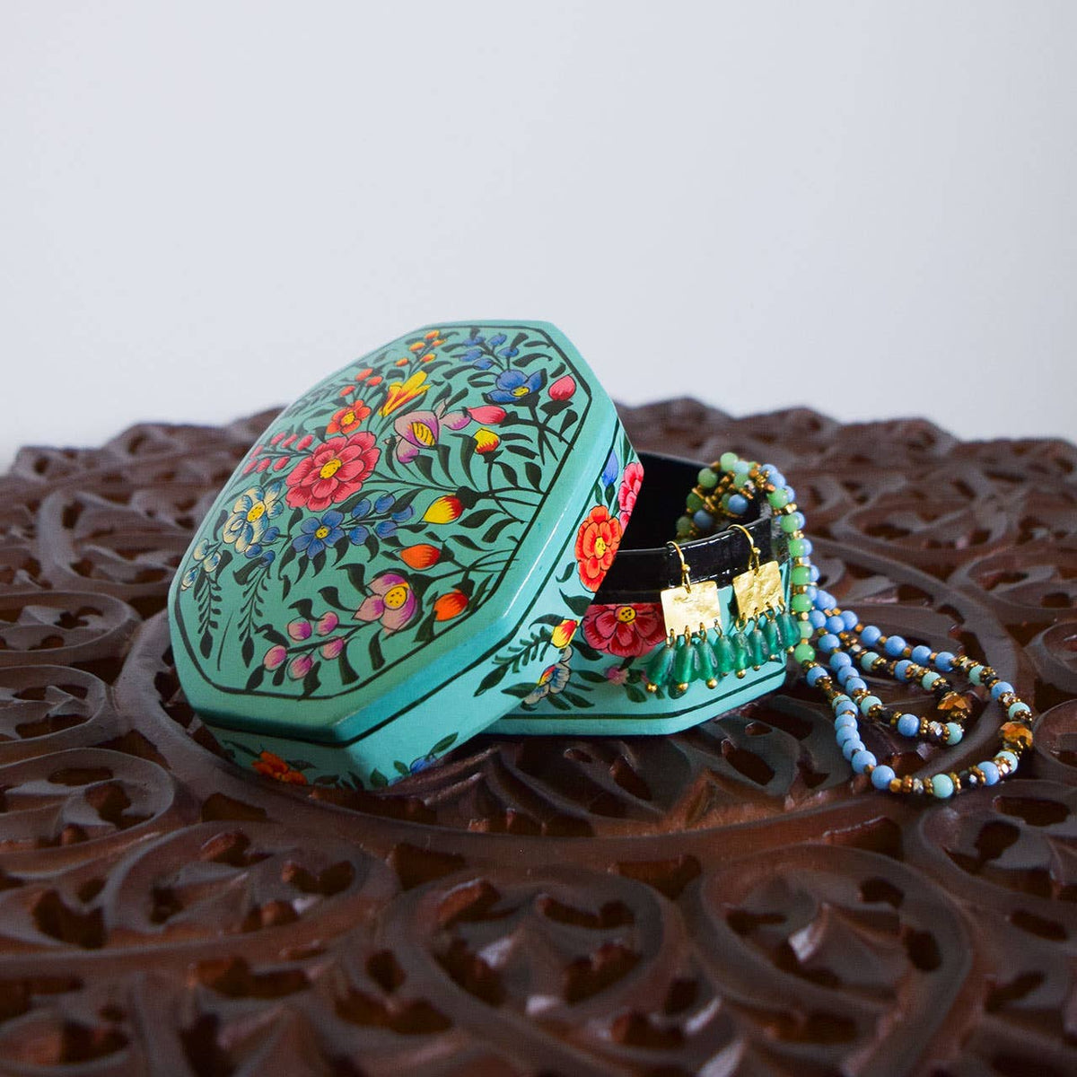 A turquoise, hexagonal Garden Handpainted Kashmiri Jewelry Box, slightly open to reveal a beaded necklace and gold-toned earrings inside.