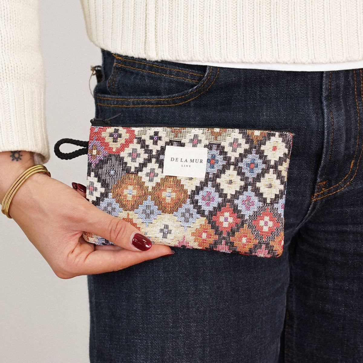 A person wearing a white sweater and dark jeans holds the Geometric Jacquard Small Makeup Bag / Coin Purse – Multicolor (Ore) from DE LA MUR LINE US Inc, featuring a patterned jacquard fabric and a white DE LA MUR label.