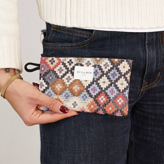 A person wearing a white sweater and dark jeans holds the Geometric Jacquard Small Makeup Bag / Coin Purse – Multicolor (Ore) from DE LA MUR LINE US Inc, featuring a patterned jacquard fabric and a white DE LA MUR label.