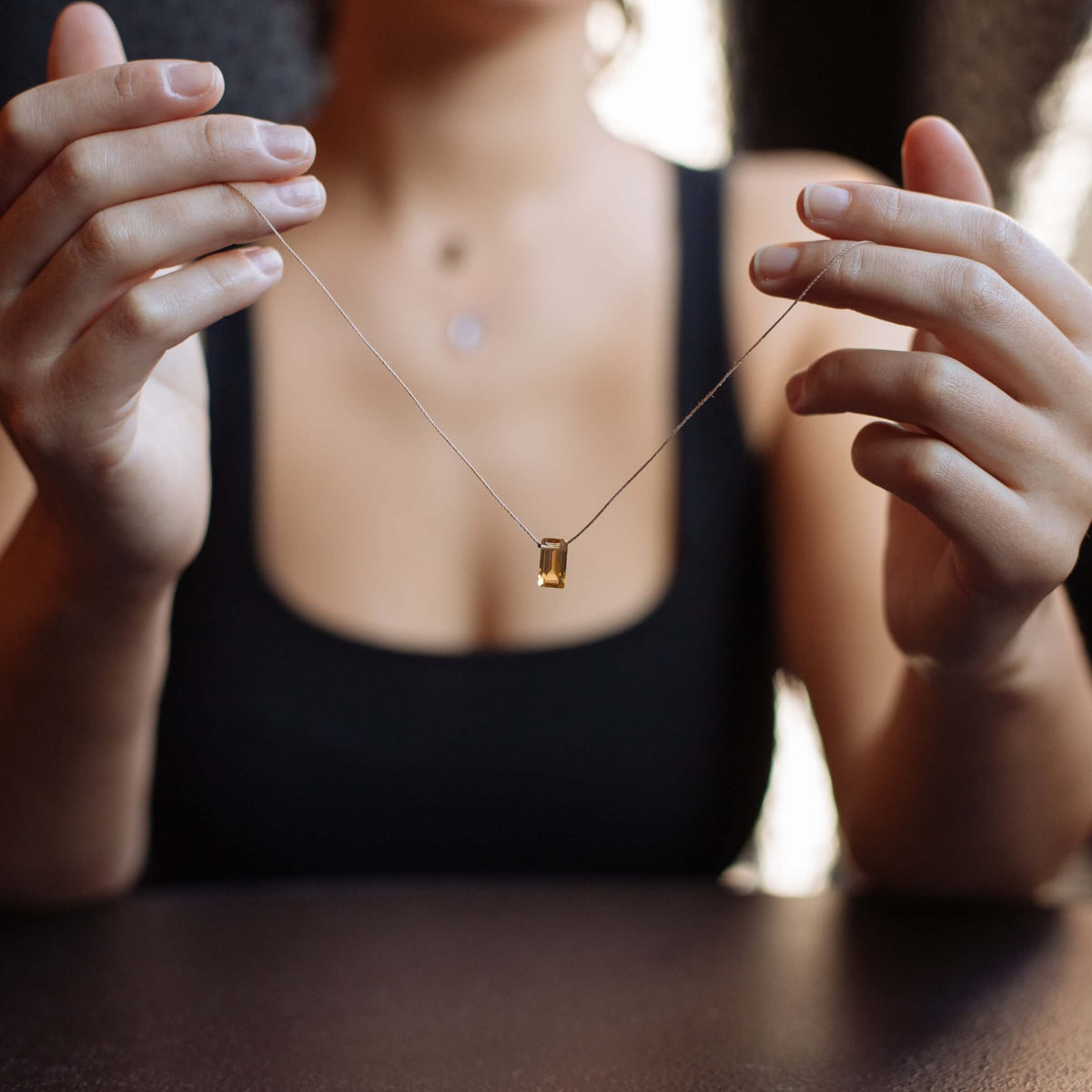A person in a black top presents the SoulKu Citrine Refined Necklace for Manifest—featuring a delicate, handmade rectangular yellow citrine pendant—displayed with both hands over a dark background.