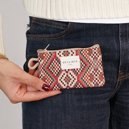 A person wearing a cream sweater and dark jeans holds the Geometric Jacquard Coin Purse – Red Multicolor (Otto) by DE LA MUR LINE US Inc in one hand, made in Barcelona.