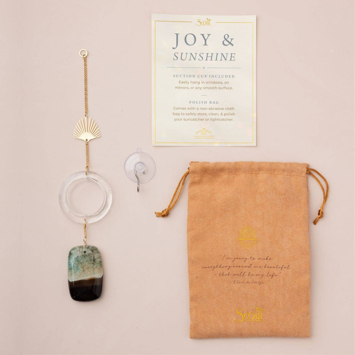 Displayed on a pale surface is the Scout Curated Wears Stone Suncatcher - Sun Rays/Black/Blue Agate, featuring a gold chain, clear ring, multicolored stones including striking blue agate, a suction cup for easy hanging, an instruction card, and a tan drawstring pouch embellished with gold text. This eye-catching piece is perfect for anyone who loves unique stone suncatchers.