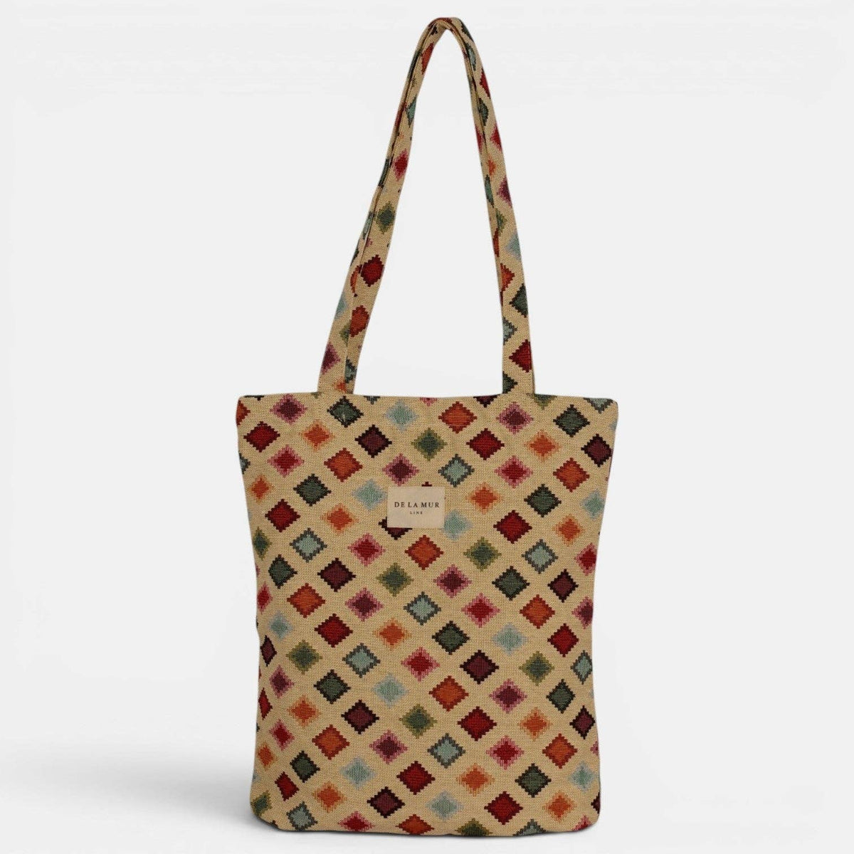 The DE LA MUR LINE US Inc Geometric Jacquard Medium Zippered Tote Bag – Multicolor (Zac) features a vibrant diamond geometric pattern in red, blue, green, and beige jacquard fabric. Designed with long handles and a small center label, this structured zippered tote stands upright against a white background.