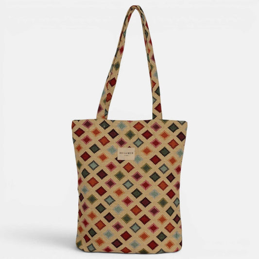 The DE LA MUR LINE US Inc Geometric Jacquard Medium Zippered Tote Bag – Multicolor (Zac) features a vibrant diamond geometric pattern in red, blue, green, and beige jacquard fabric. Designed with long handles and a small center label, this structured zippered tote stands upright against a white background.