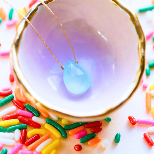 The Chalcedony Luxe Necklace for Happy Birthday by SoulKu—a teardrop-shaped blue gemstone—rests in a white, gold-rimmed bowl with colorful sprinkles, making it a unique handmade gift.