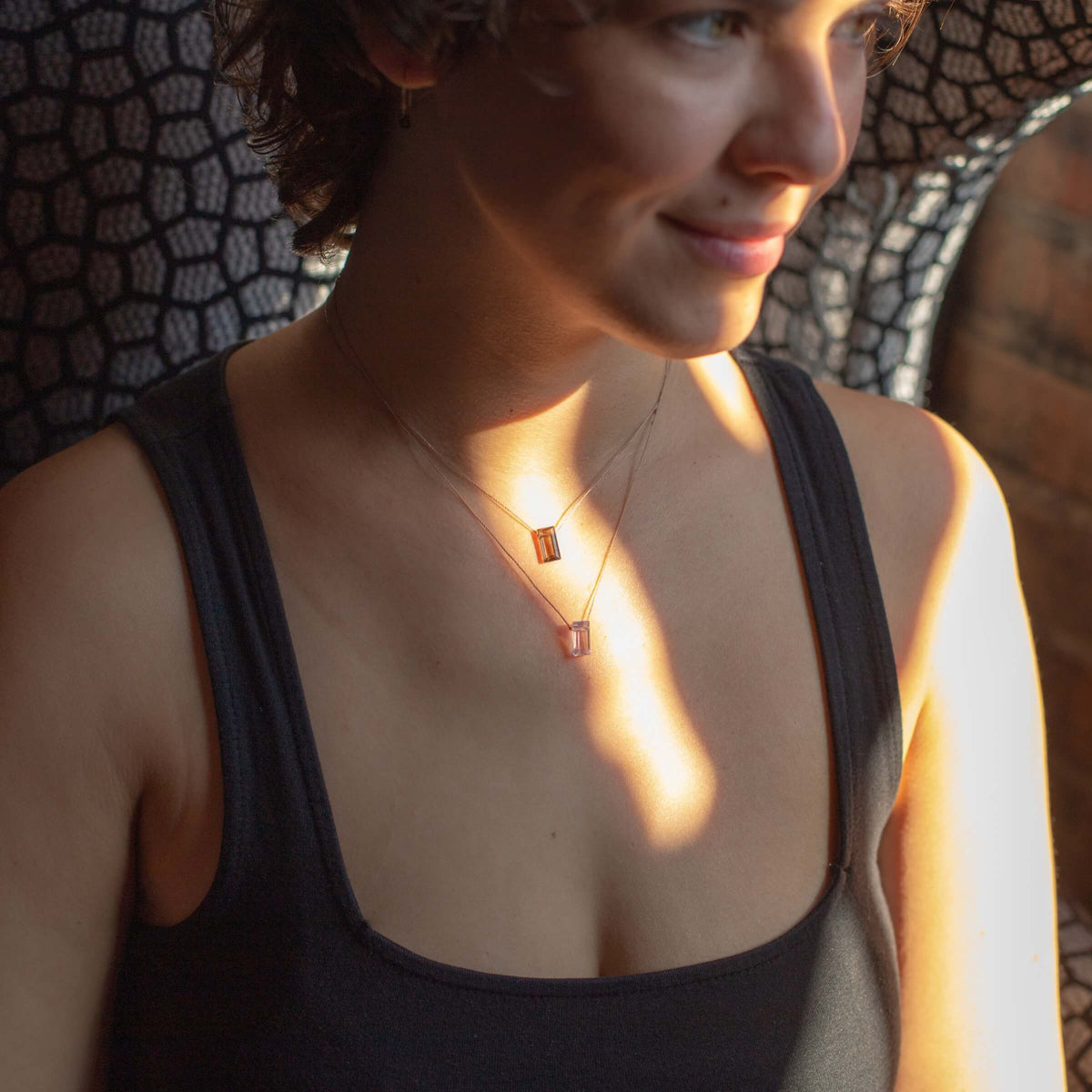 A person in a sleeveless black top sits in dappled sunlight, smiling gently and looking to the side while wearing two SoulKu Amethyst Refined Necklace for Fearless with rectangular gemstone pendants.