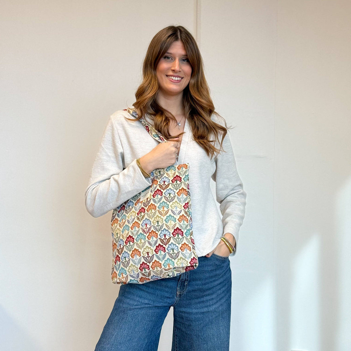A woman with long brown hair smiles while wearing a cream sweater and blue jeans, holding the DE LA MUR LINE US Inc Floral Jacquard Medium Zippered Tote Bag – Multicolor (Flora) on her shoulder, standing against a plain light-colored wall.