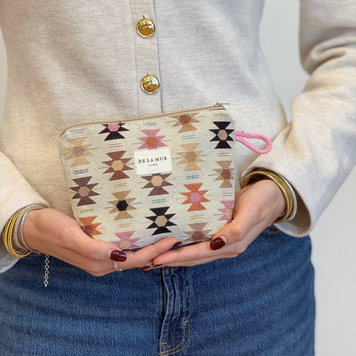 A person in a cream cardigan and blue jeans holds the Geometric Jacquard Small Makeup Bag – Pink & Orange (Royal) by DE LA MUR LINE US Inc. The jacquard fabric pouch displays vibrant pink, orange, and black Aztec-inspired geometric patterns and features a soft pink loop.