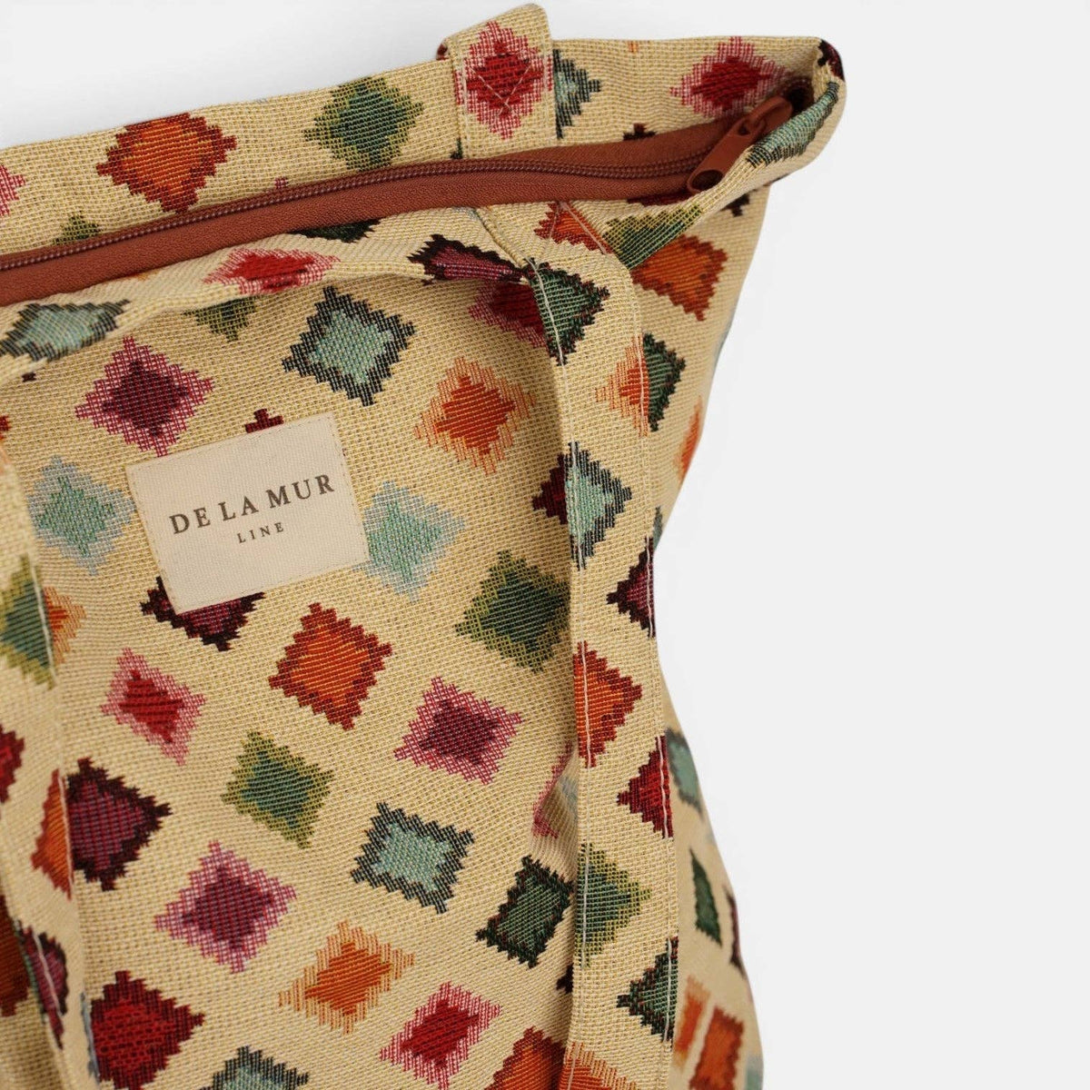 The Geometric Jacquard Medium Zippered Tote Bag – Multicolor (Zac) by DE LA MUR LINE US Inc is crafted from jacquard fabric with a vibrant multicolor diamond pattern. It features a brown zipper and a front label displaying the brand’s name. The tote bag is shown against a white background.