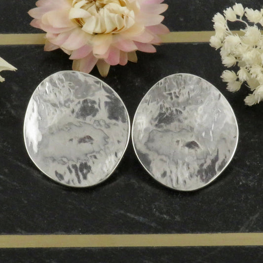 Two oval, hammered silver clip-on earrings from Danon are displayed on a dark marble surface with cream and pink dried flowers in the background.